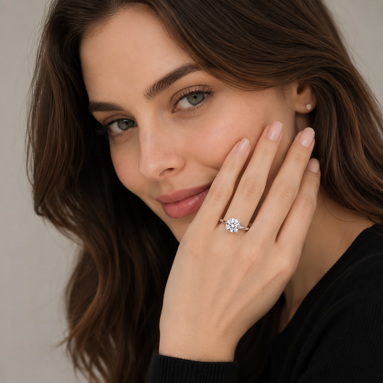 A close-up lifestyle shot of a woman with brown hair wearing a diamond engagement ring on her finger, resting her hand on her cheek against a neutral beige background.