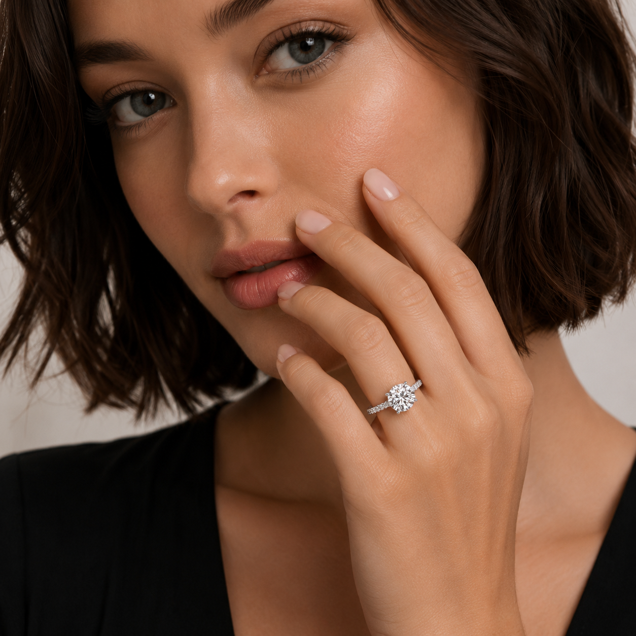 A close-up lifestyle shot of a woman wearing a sparkling diamond engagement ring on her finger, with her hand resting near her face.