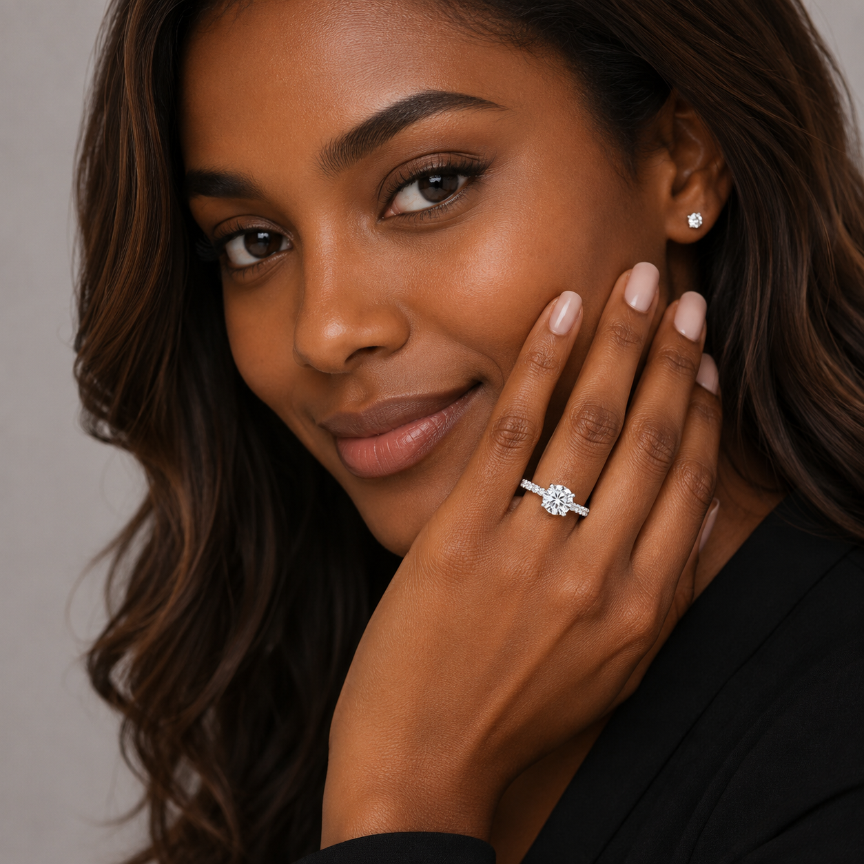 A close-up lifestyle shot of a woman wearing a sparkling diamond engagement ring on her finger, with a small matching diamond stud earring on her ear. The ring features a round brilliant-cut center stone set in a four-stone diamond band.
