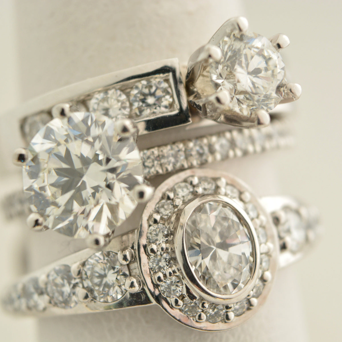 A close-up of a stack of silver-toned rings with multiple round and oval diamonds, displayed on a soft cream ring stand against a light neutral background.