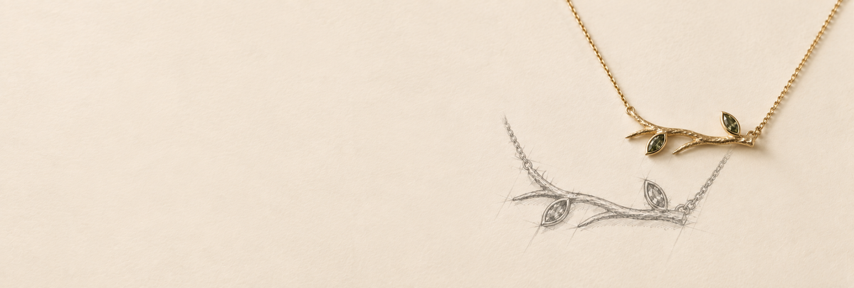 A delicate necklace with a gold-toned branch and leaf pendant featuring small green stone accents, displayed on a warm cream background next to a matching silver-toned branch necklace.