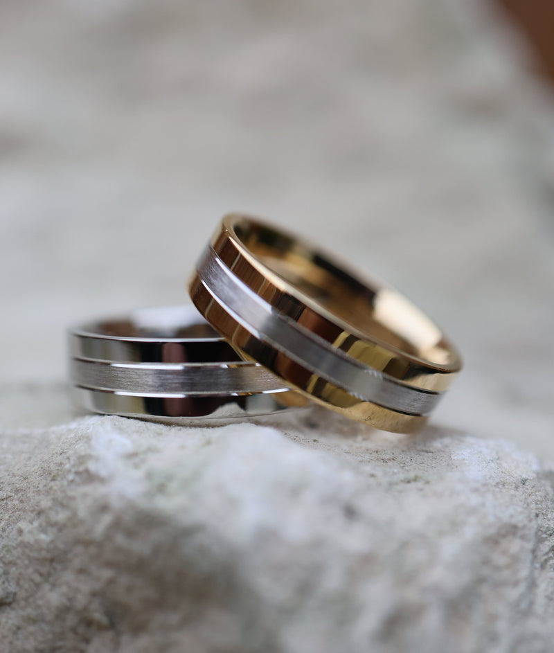 Two slim metal bands rings resting on a textured light gray stone surface. One ring is polished silver-toned with a central gold inlay, and the other is polished gold with a central silver inlay, creating a two-tone effect.