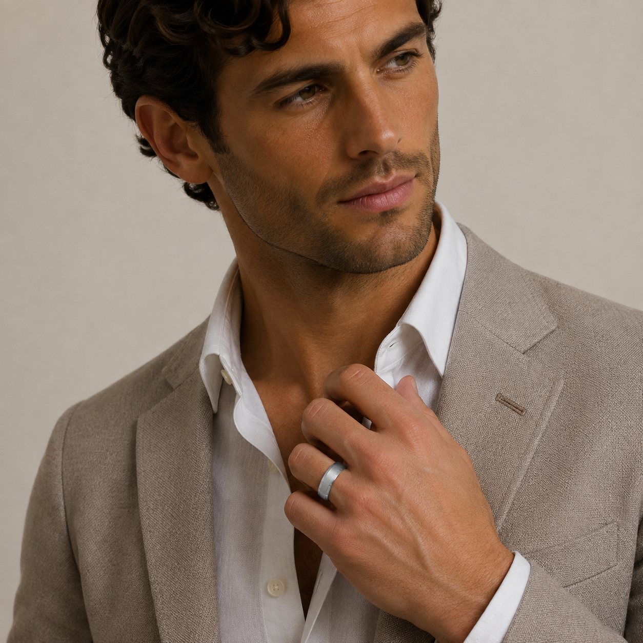 A close-up of a man wearing a light gray blazer and white dress shirt, adjusting his collar while wearing a simple silver band ring on his finger, set against a neutral beige background.