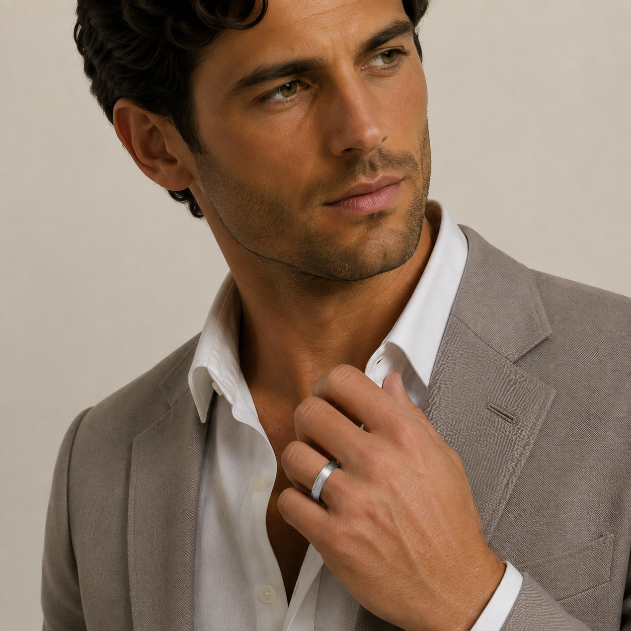 A close-up of a man wearing a light gray blazer and white dress shirt, adjusting his collar and wearing a simple silver band ring on his finger, set against a plain beige background.