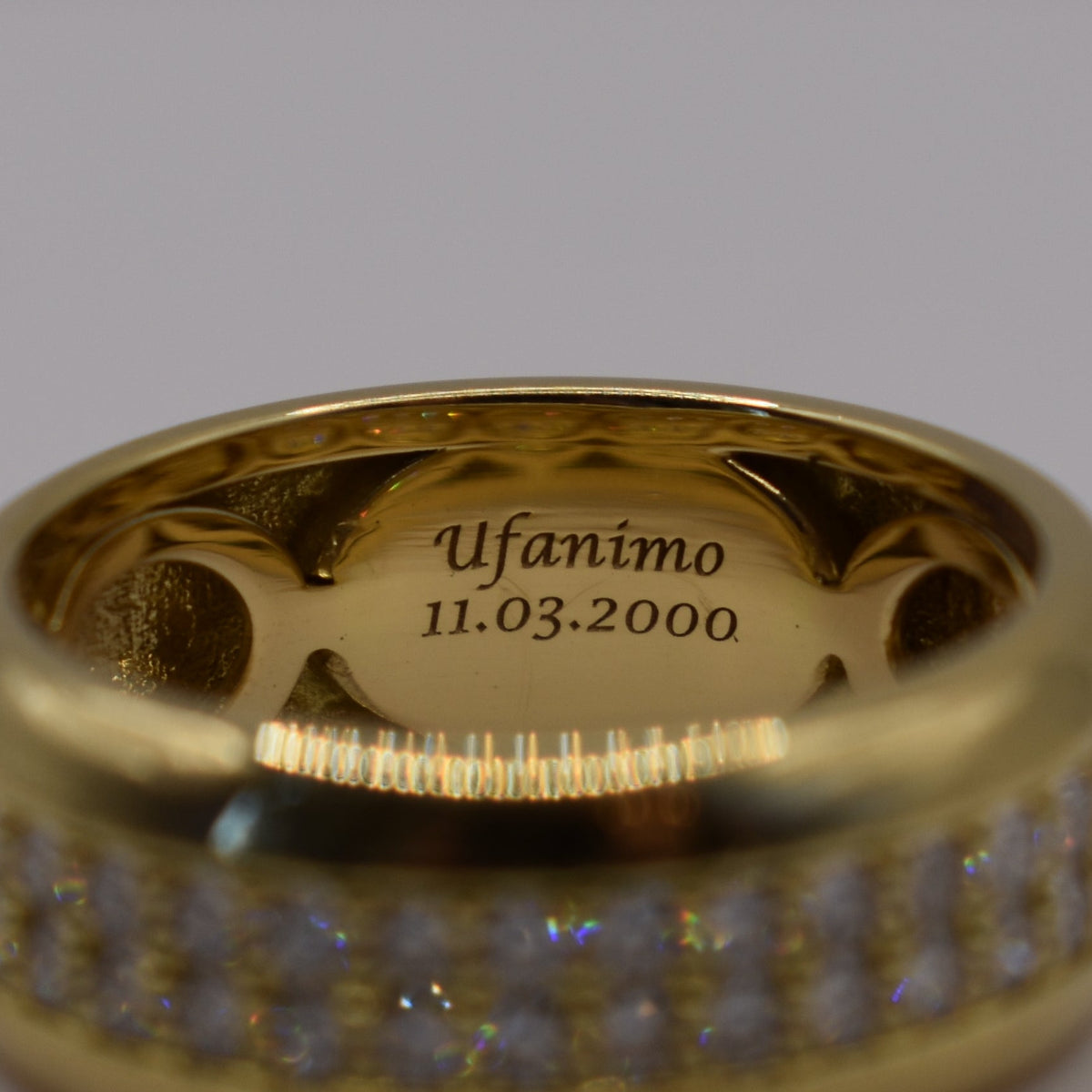 A gold band ring with a row of small clear stones on the front, photographed on a plain light gray background. The inside of the ring is engraved with the name 'Ufanimo' and the date '11.
