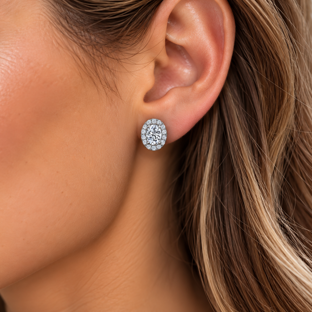 A close-up of a woman's ear wearing a small oval stud earring with a halo of clear stones in a silver-toned setting.