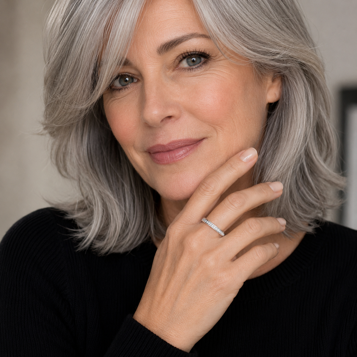A close-up portrait of a woman with shoulder-length gray hair wearing a slim silver band ring on her finger, posed against a soft neutral background.