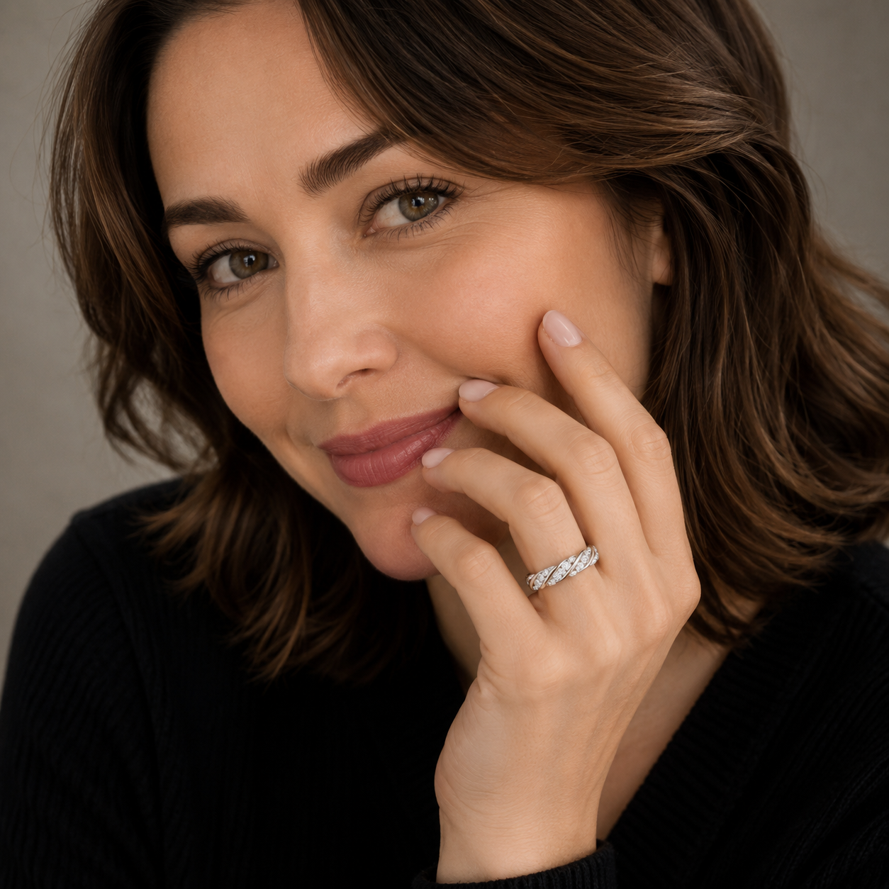 A close-up portrait of a woman with brown hair wearing a silver band ring with a textured, diamond-like pattern on her ring finger, set against a soft gray background.