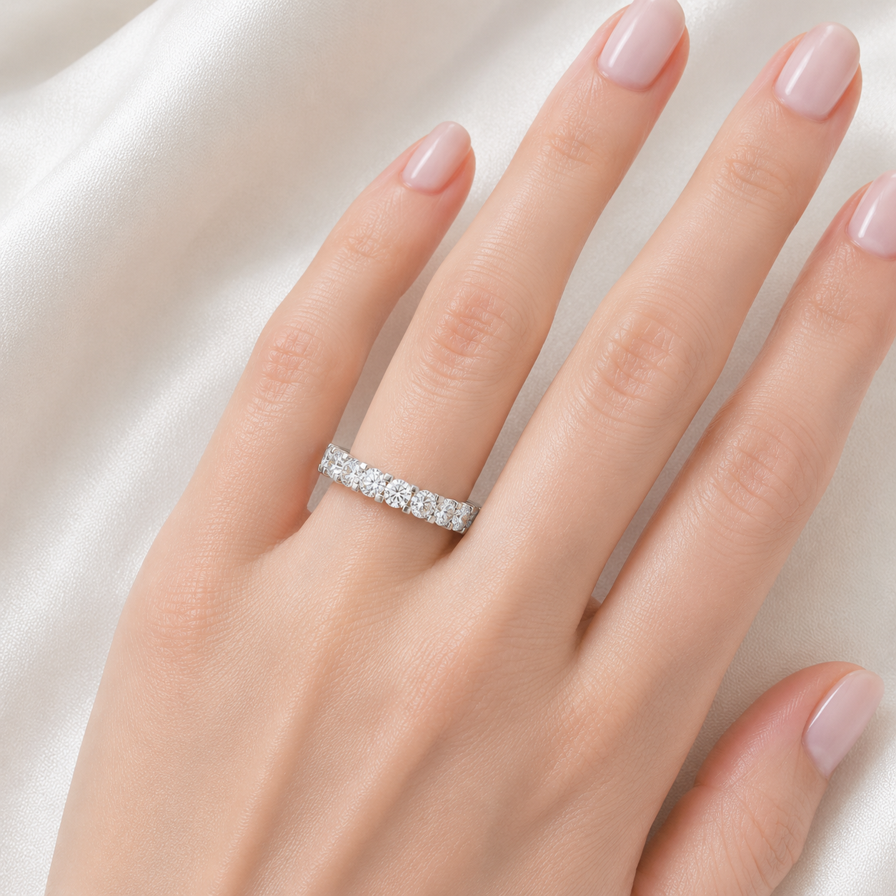 A close-up of a hand with pale pink manicured nails wearing a silver eternity ring set with small clear stones, resting against a soft white satin fabric background.