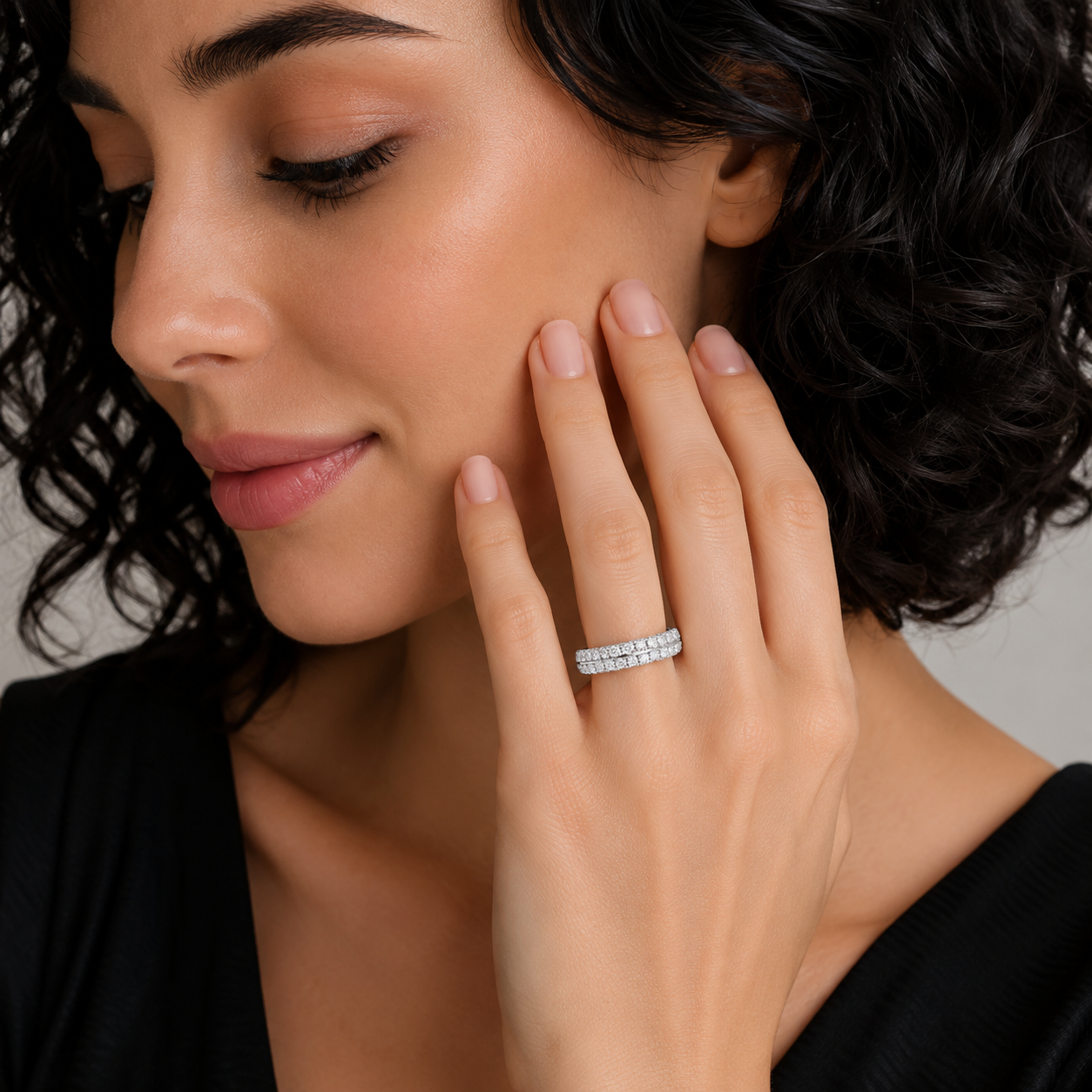 A close-up lifestyle shot of a woman with dark curly hair wearing a slim silver band ring set with small clear stones on her ring finger.