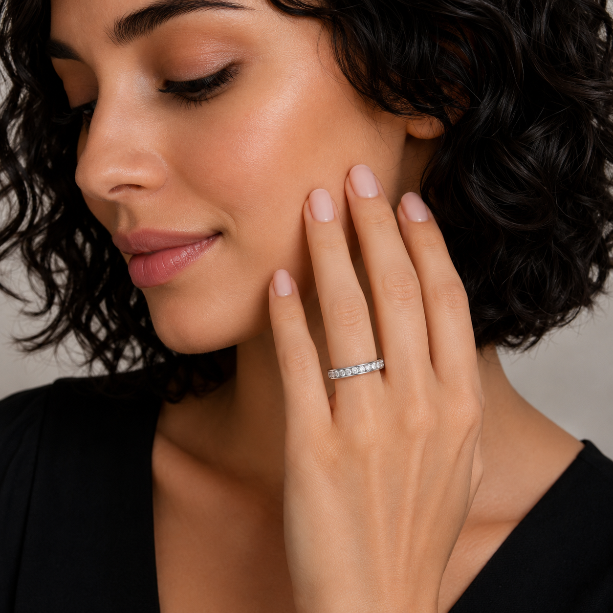 A close-up lifestyle shot of a woman wearing a thin silver diamond eternity ring on her finger, with her hand resting on her cheek against a neutral gray background.