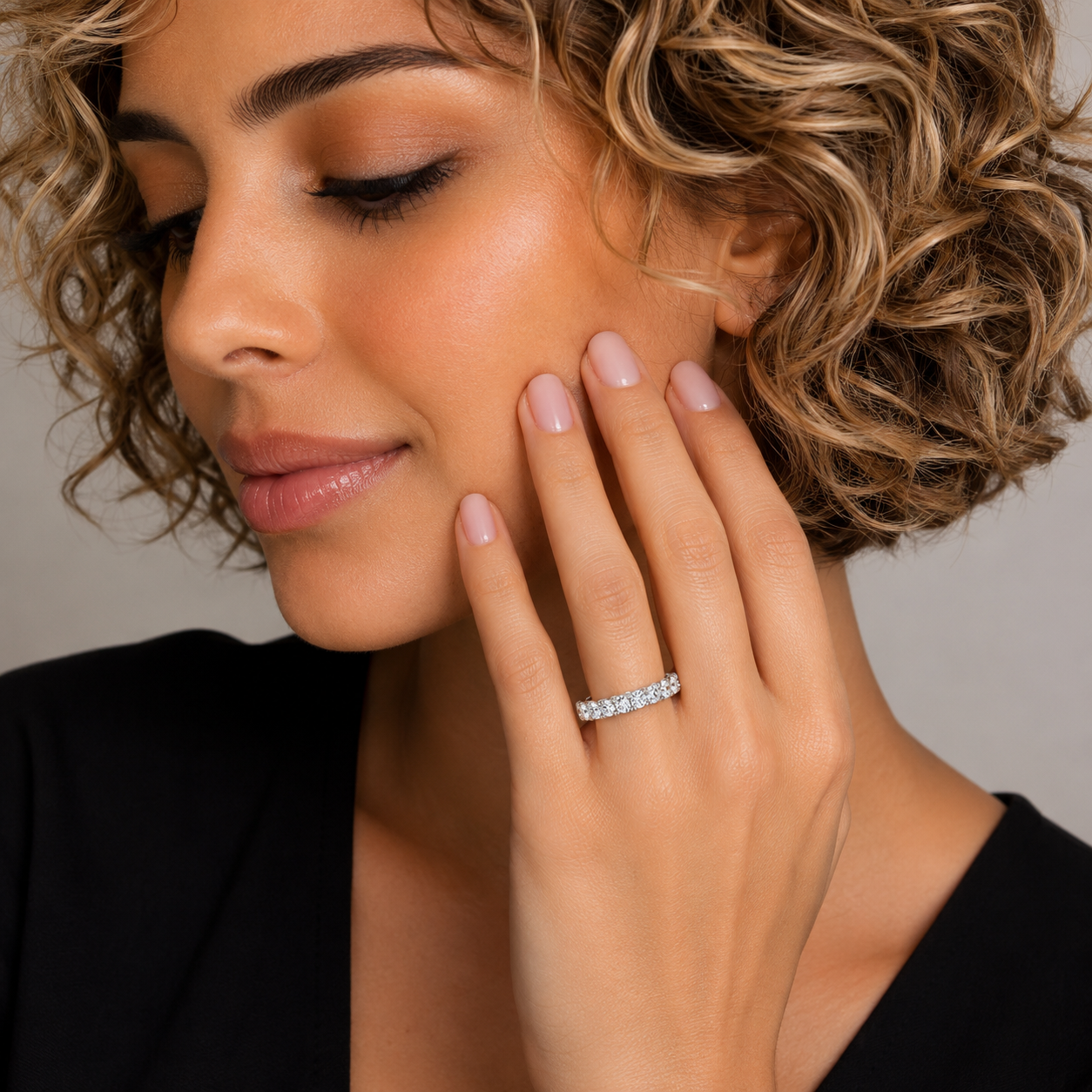 A close-up lifestyle shot of a woman with short curly blonde hair wearing a silver diamond eternity ring on her finger, with her hand resting near her face.
