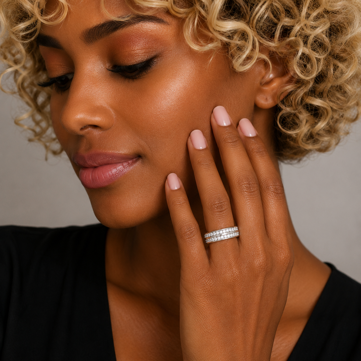 A close-up lifestyle shot of a woman with curly blonde hair wearing a delicate silver band ring set with small clear stones on her ring finger.