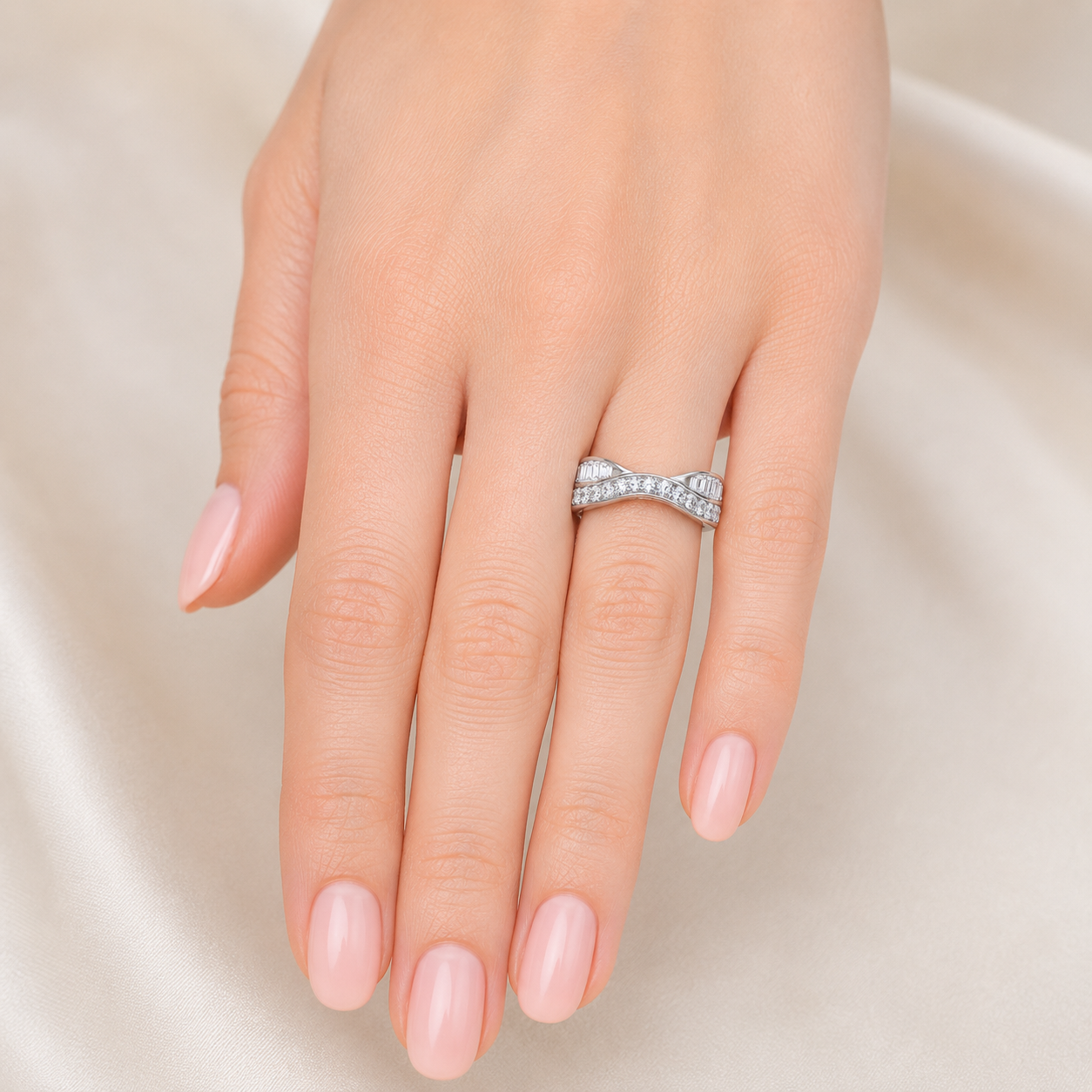 A close-up of a hand with pale pink manicured nails wearing a silver ring set with small clear stones in a bow-like twisted design, against a soft cream satin background.