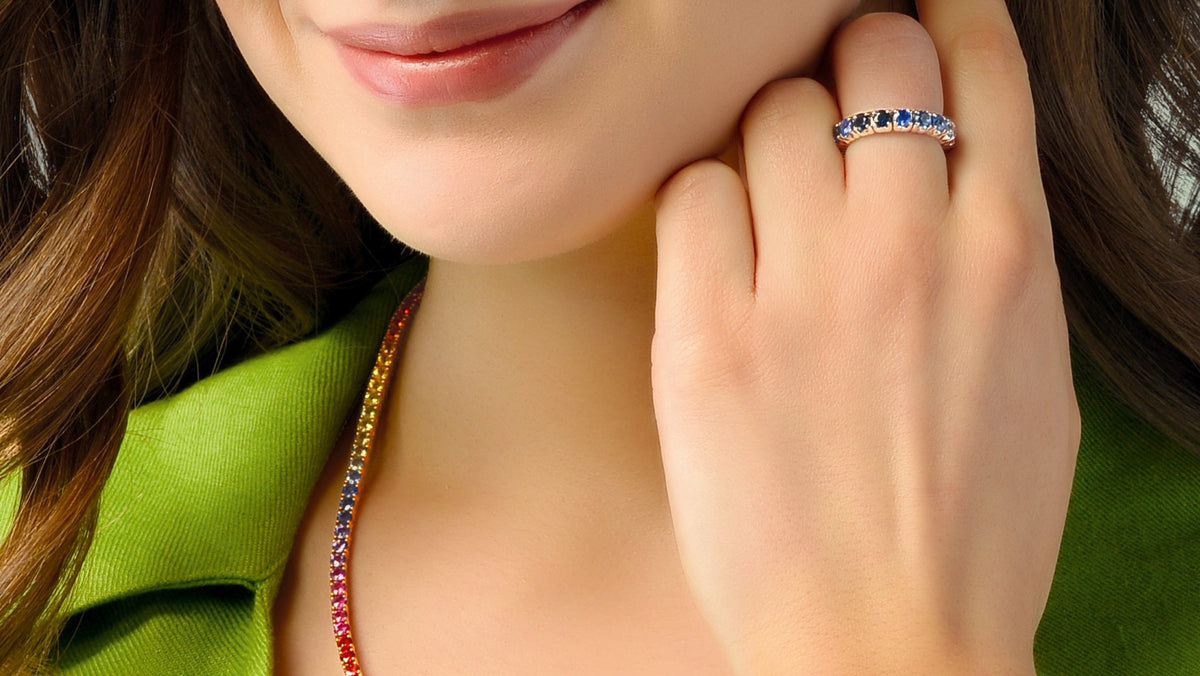 A close-up lifestyle shot of a woman wearing a delicate, multi-colored beaded necklace and a matching beaded ring on her finger. The jewelry features small, colorful gemstones in a rainbow sequence.