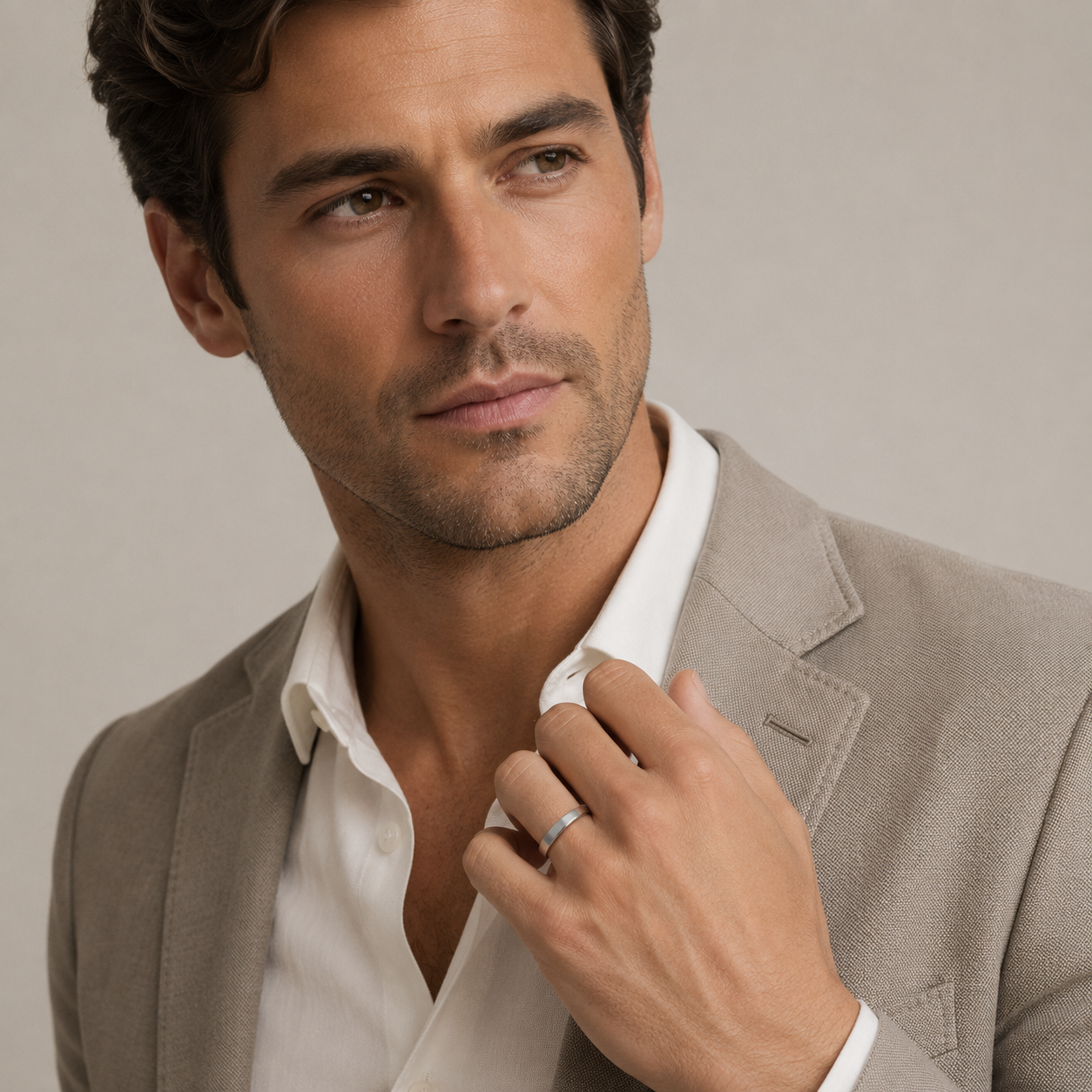 A close-up of a man wearing a light beige blazer over an open white dress shirt, with his hand adjusting the collar and a slim silver band visible on his ring finger, against a soft beige studio background.