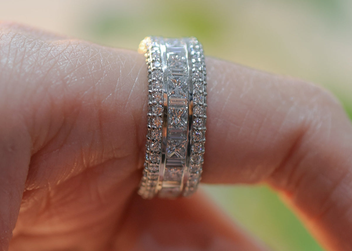 A close-up of a silver-toned double band ring with pavé-set clear stones worn on a finger. The ring features a central rectangular block design with smaller stones around it.