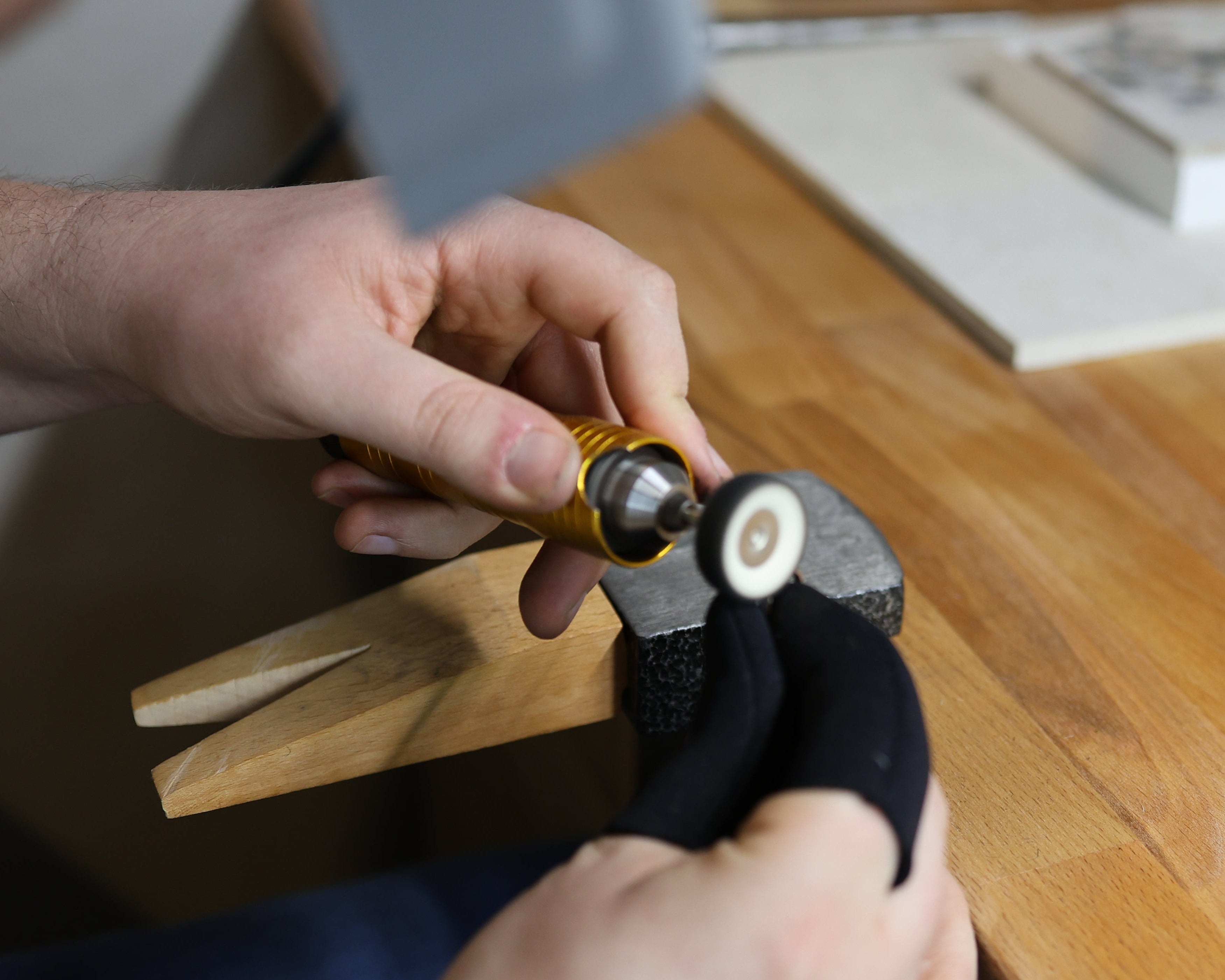 A close-up lifestyle shot of a person's hands using a small rotary tool with a grinding wheel attachment to work on a black sock held in a wooden clamp on a wooden workbench.