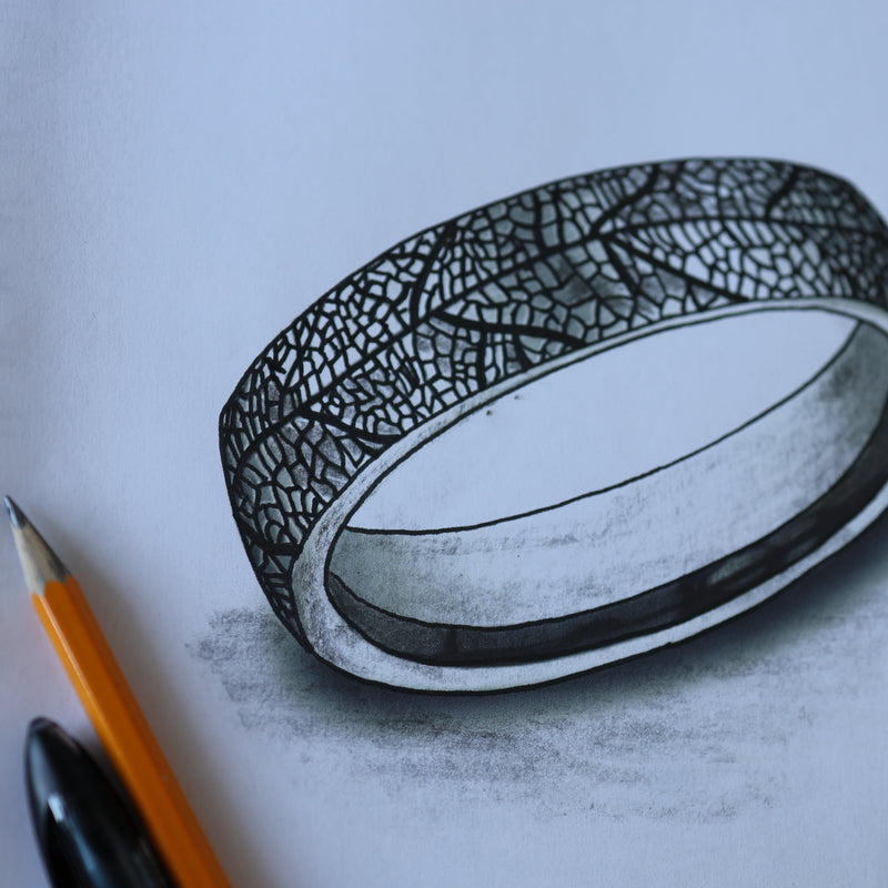 A realistic pencil sketch of a textured silver ring with a leaf-like pattern, shown on a white sheet of paper next to an orange pencil and a black pen.