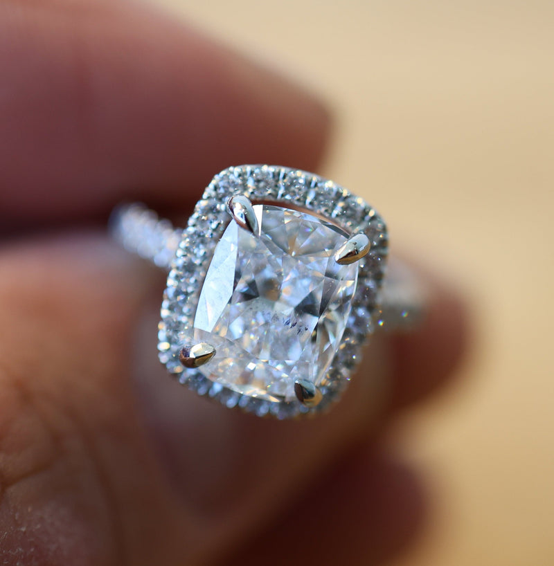 A close-up shot of a cushion-cut diamond halo ring on a person's finger, set against a warm, blurred wooden background.