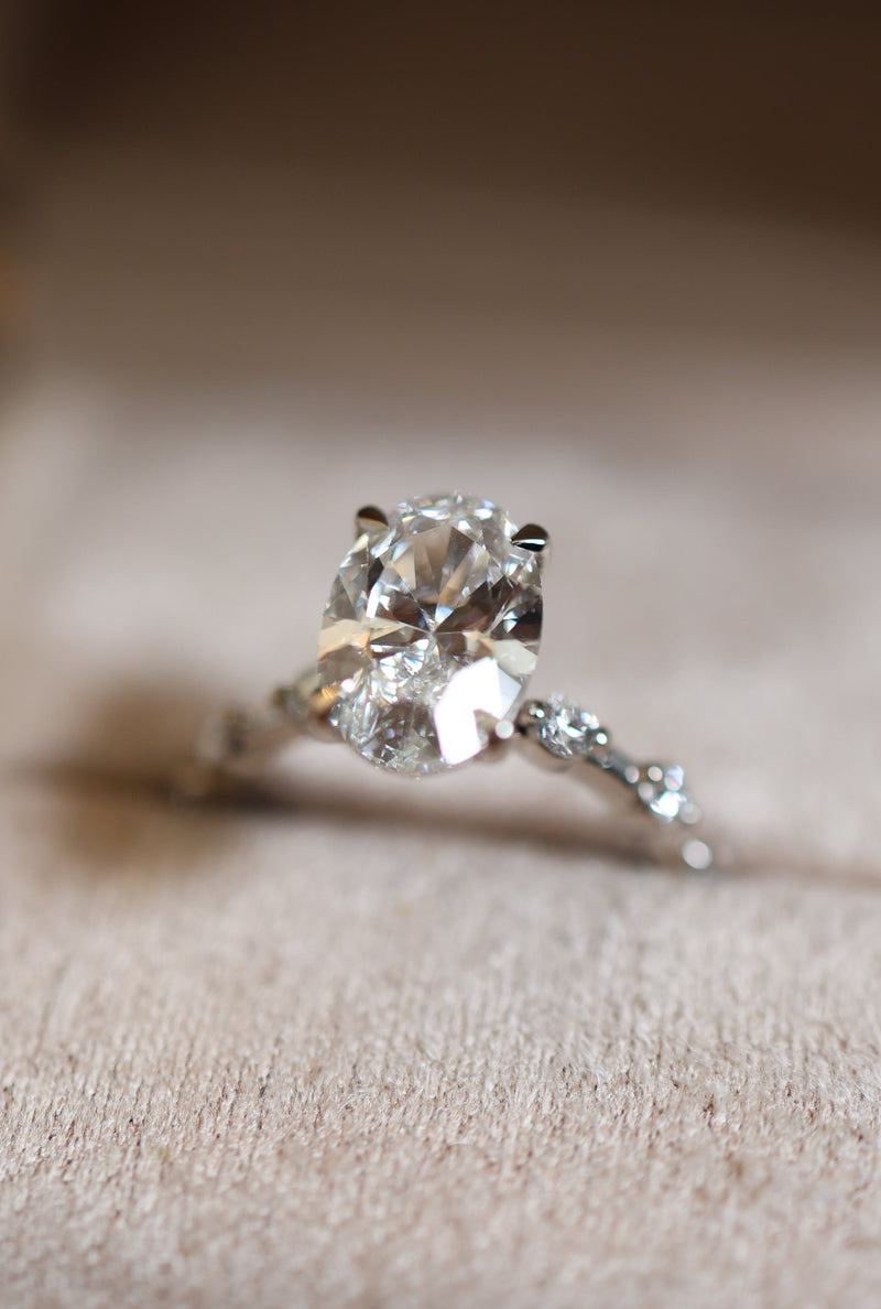 A silver solitaire engagement ring with a large round brilliant-cut center stone and a smaller accent stone on the band, resting on a beige textured ring box.