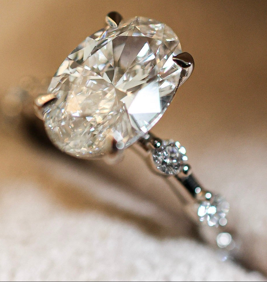 An oval-cut diamond engagement ring with a silver-toned band featuring small accent diamonds, resting on a soft beige textured surface.