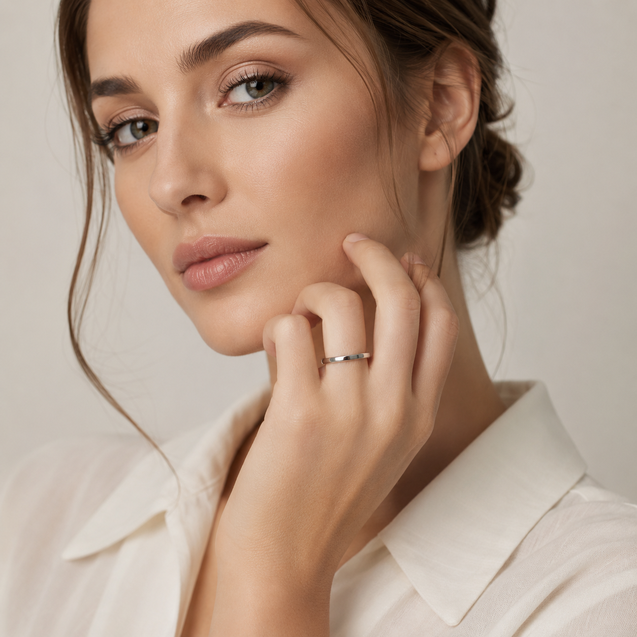 A close-up of a woman wearing a thin silver band ring on her finger, posing with her hand near her face against a soft, light beige background.