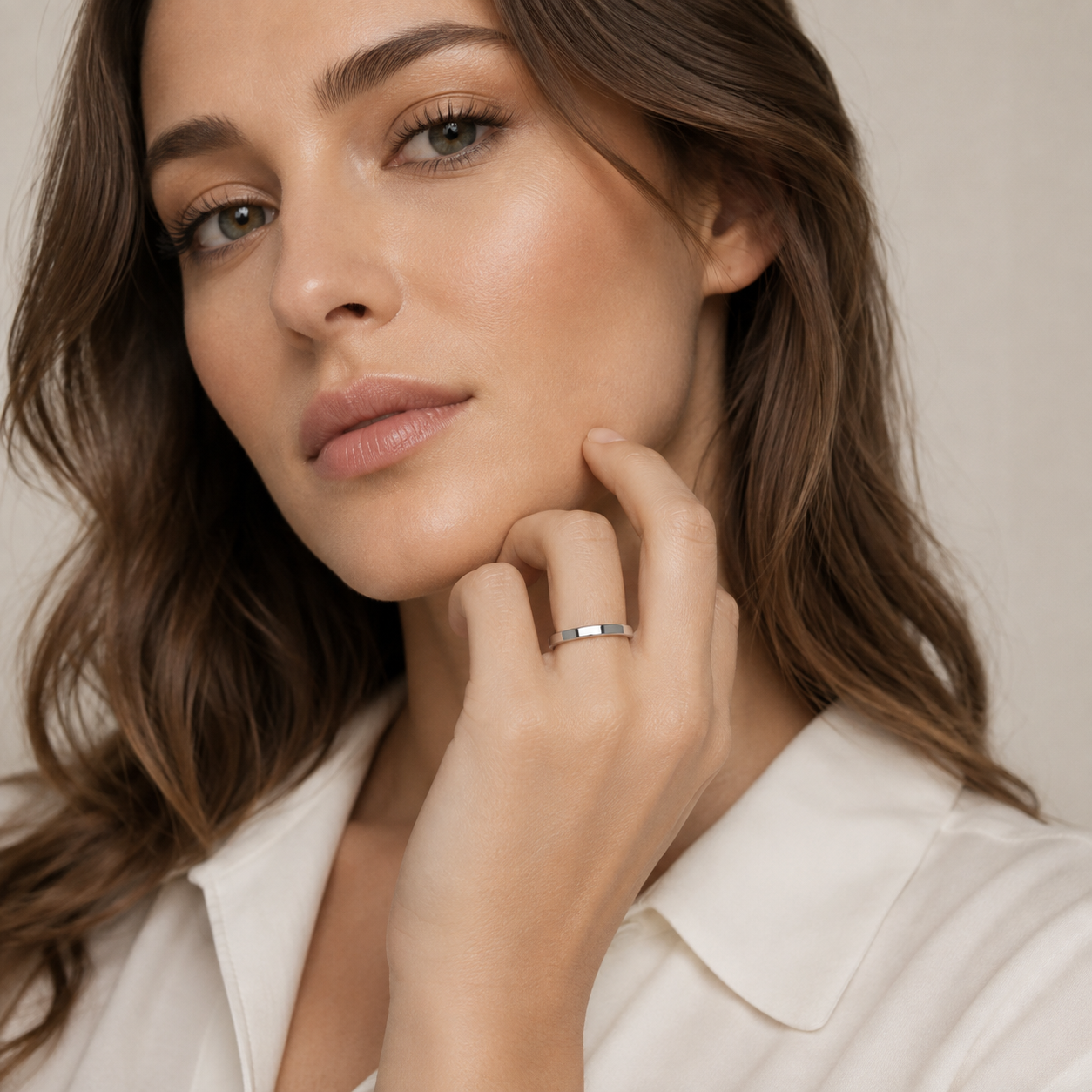 A close-up of a woman wearing a slim silver band ring with a small clear center stone, styled with a white collared shirt against a soft beige background.