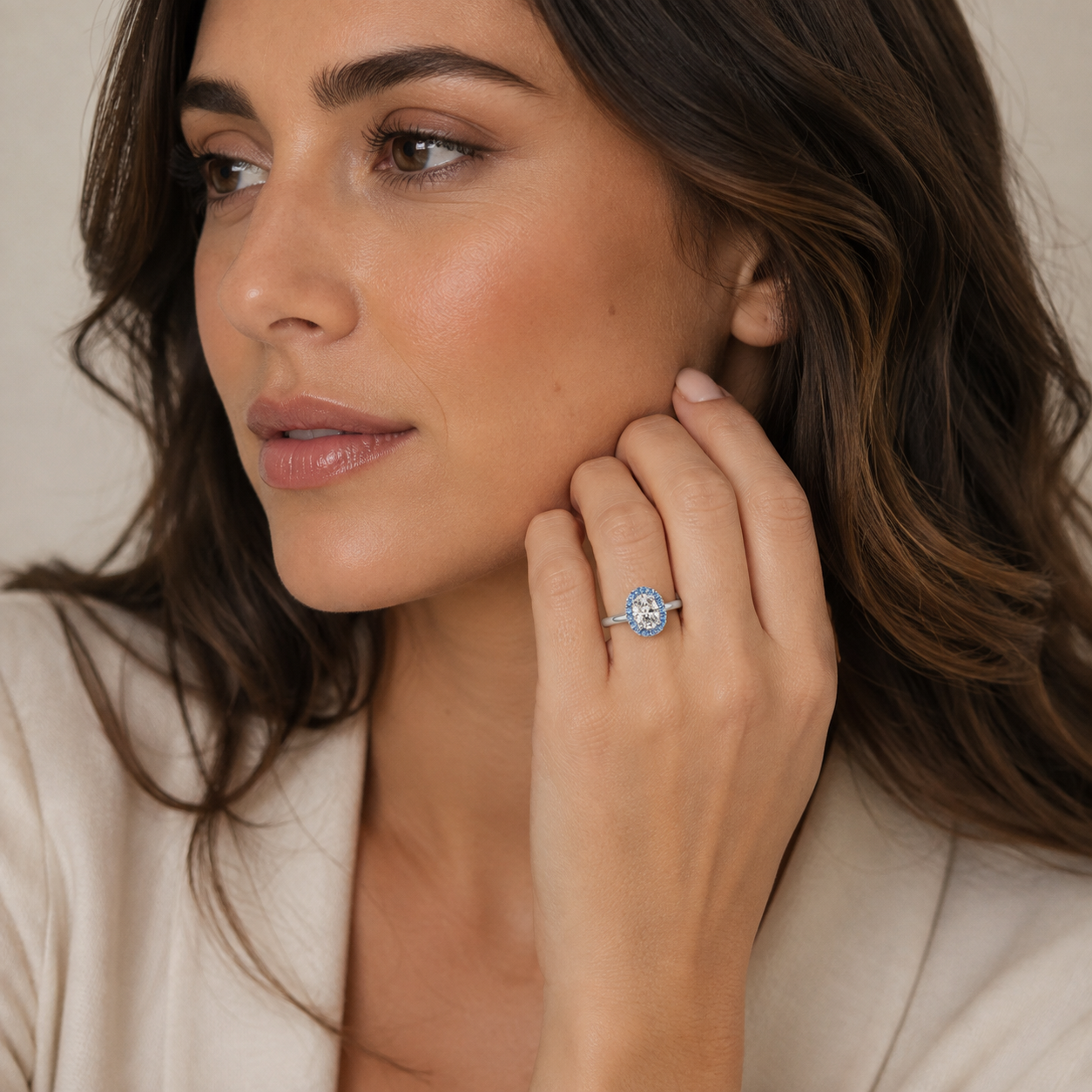 A close-up lifestyle shot of a woman wearing a silver ring with a round-cut blue gemstone on her finger, set against a soft beige background.