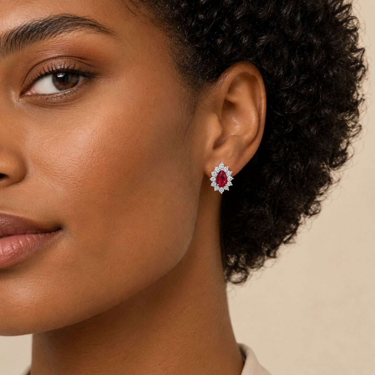 A close-up of a model's ear wearing a single teardrop-shaped stud earring with a central red gemstone surrounded by small clear stones in a silver-toned setting, against a plain beige background.