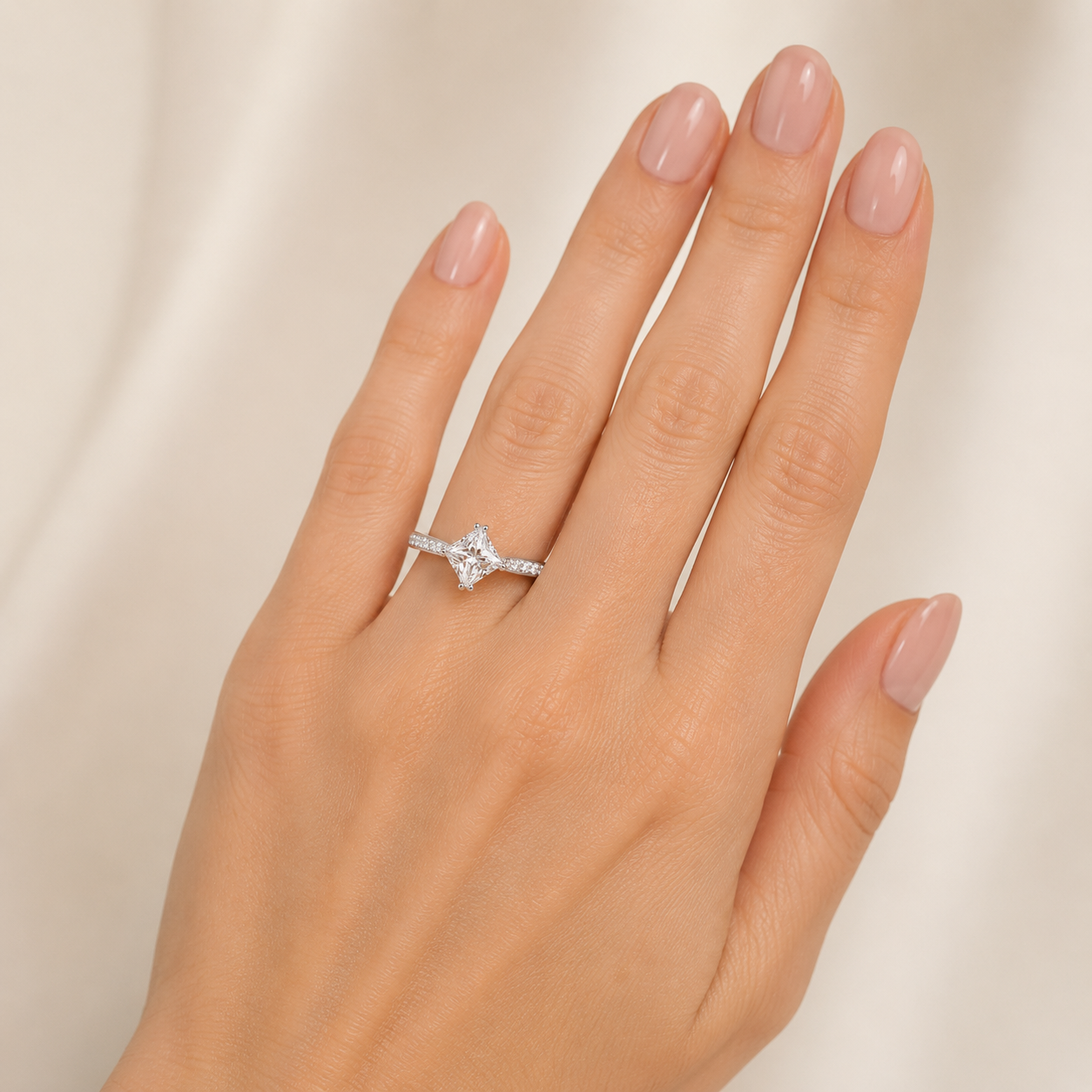 A close-up of a hand wearing a delicate silver-toned ring with a central clear gemstone, set against a soft cream-colored fabric background.