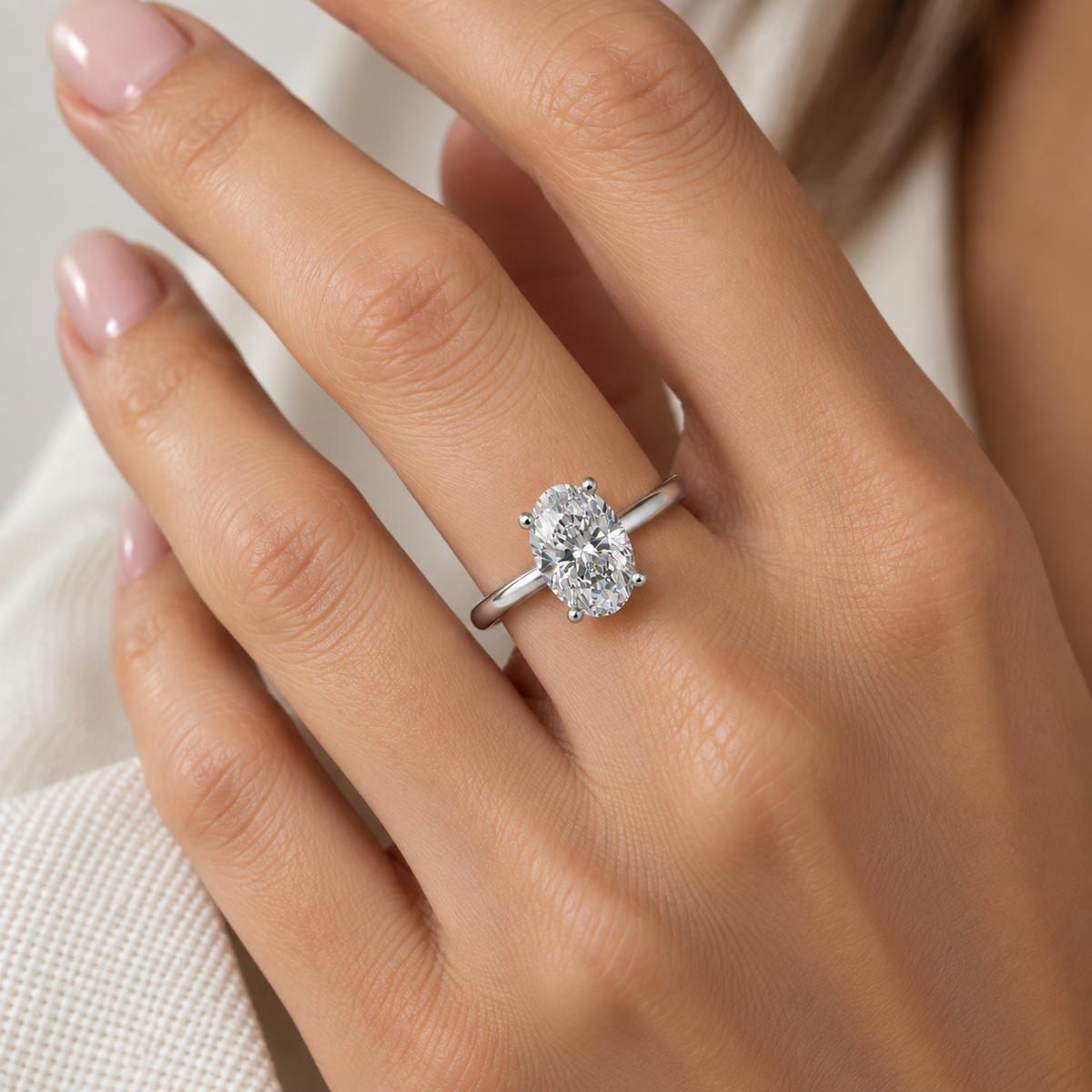 A close-up of a hand wearing a silver engagement ring with a round brilliant-cut diamond. The ring is shown on the ring finger against a soft, light beige background.