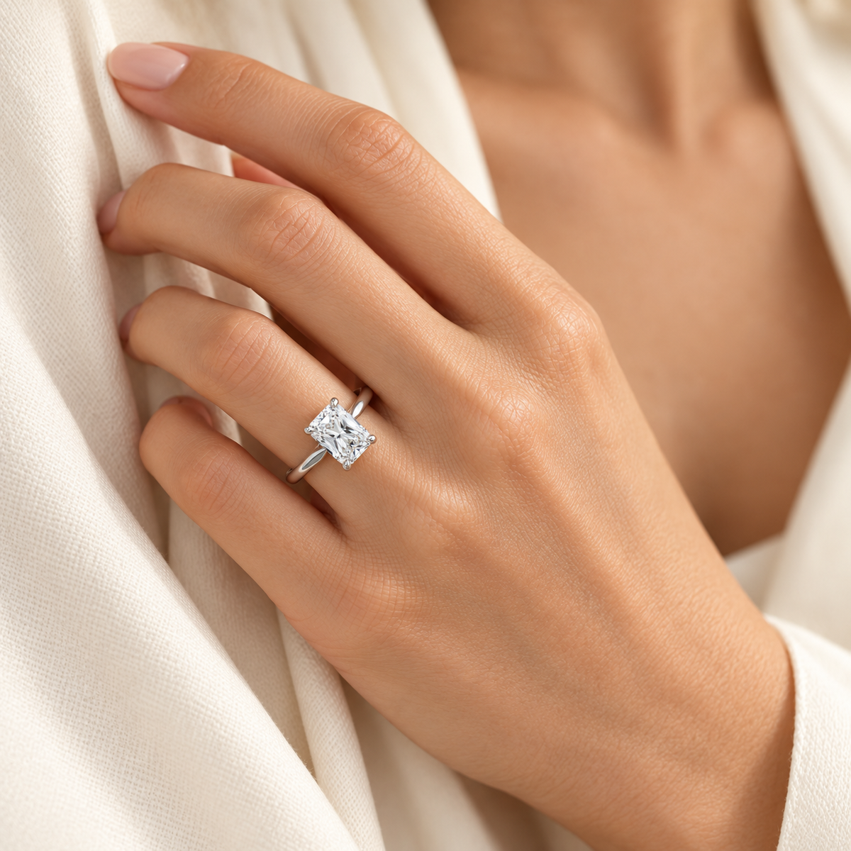 A close-up of a hand wearing a silver engagement ring with a radiant clear diamond, set against a soft cream-colored fabric background.