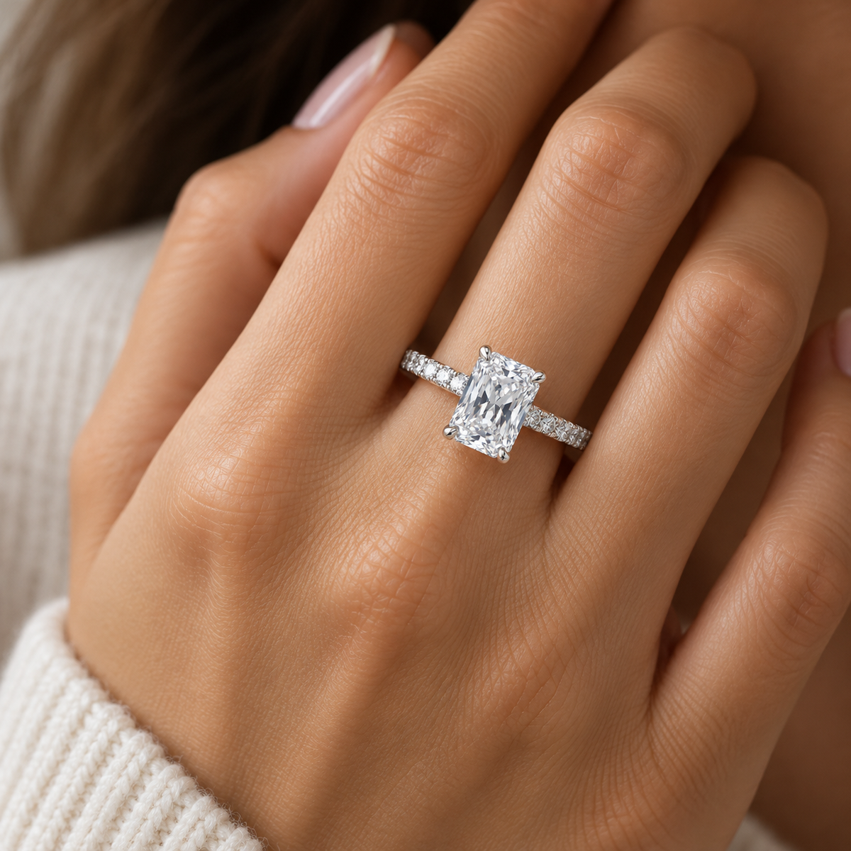 A close-up of a woman's hand wearing a silver engagement ring with a rectangular clear gemstone and a pavé band.
