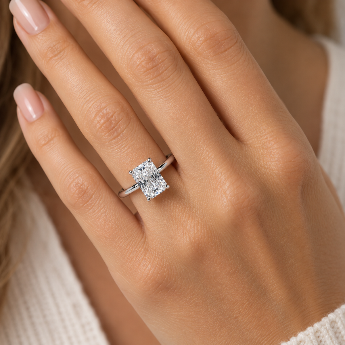A close-up of a person's hand wearing a silver-toned ring with a rectangular clear gemstone, set against a soft cream knit top.