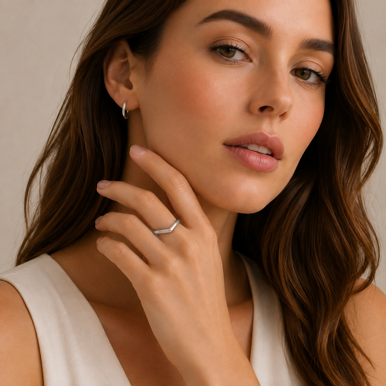 A close-up of a woman wearing a thin silver band ring on her finger and a small silver hoop earring in her ear, against a soft beige background.