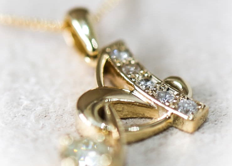 A gold pendant necklace featuring a stylized abstract design with small clear stones and a dangling round clear stone, shown in a close-up shot on a light textured surface.