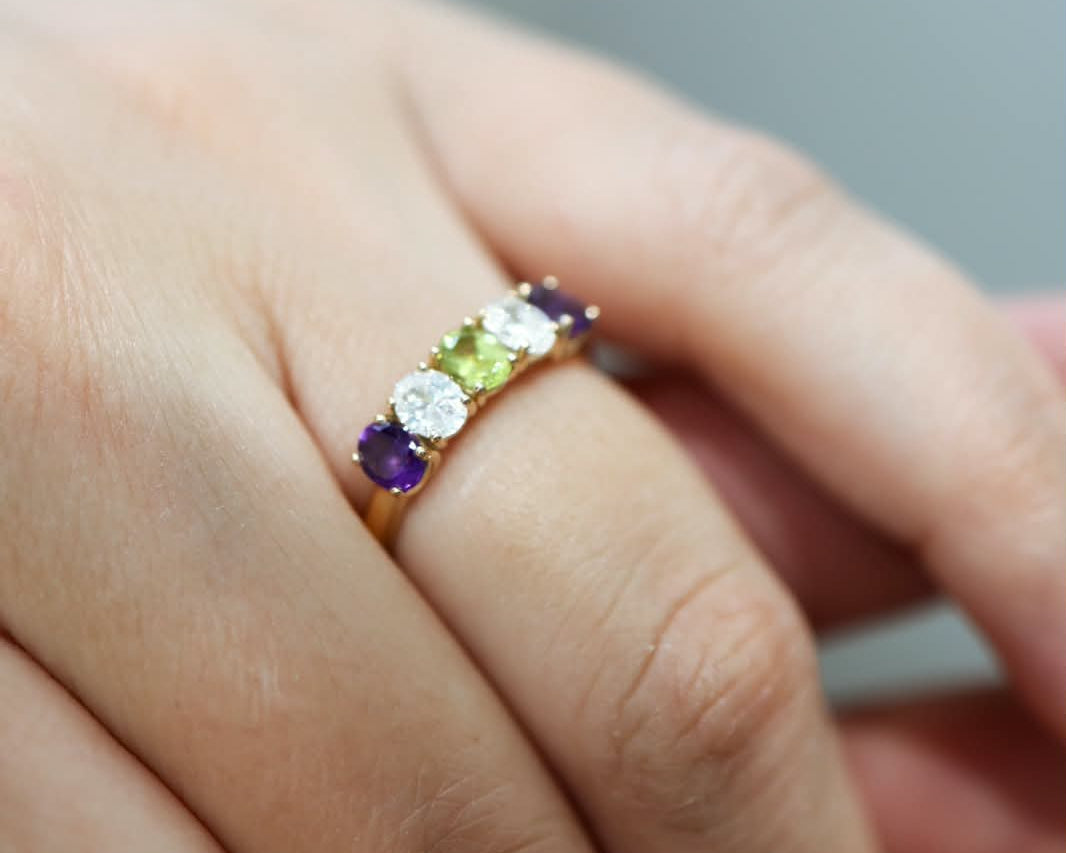 A close-up of a hand wearing a gold band ring set with four round gemstones in purple, white, green, and dark purple.
