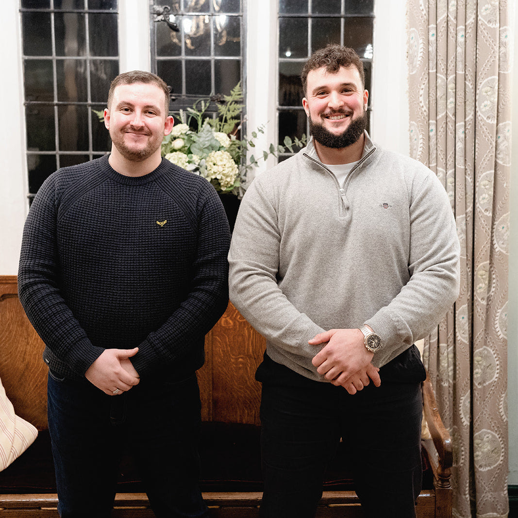 Two men standing indoors in front of a window and curtains, wearing quarter-zip sweaters. The man on the left wears a black waffle-knit sweater with a small yellow embroidered logo, and the man on the right wears a light gray quarter-zip pullover.