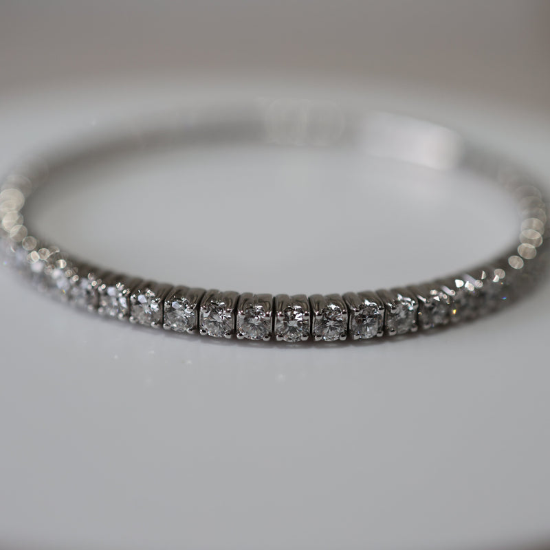 A close-up of a diamond tennis bracelet in a silver-toned setting, resting on a light gray surface.