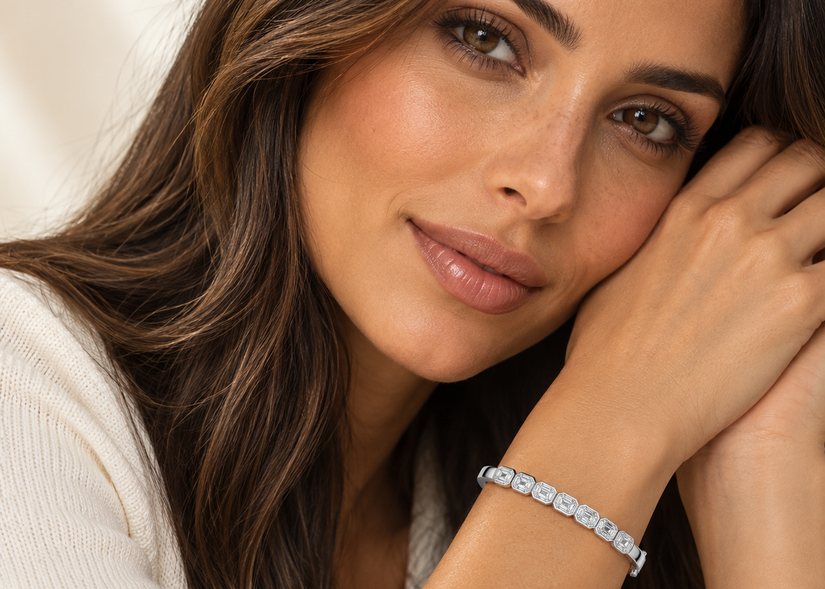 A close-up lifestyle shot of a woman with long brown hair wearing a silver tennis bracelet with clear round-cut stones. She is wearing a white knit top and resting her head on her hand, with the bracelet clearly visible on her wrist.