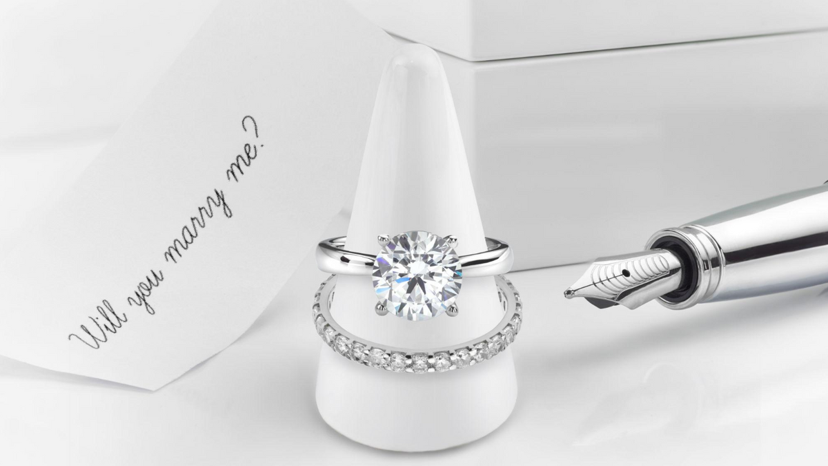 A silver engagement ring with a large round clear center stone is displayed on a white ring cone, with a matching silver band featuring small clear stones beside it. A white card reading 'Will you marry me?' and part of a silver fountain pen are also visible on a clean white background.