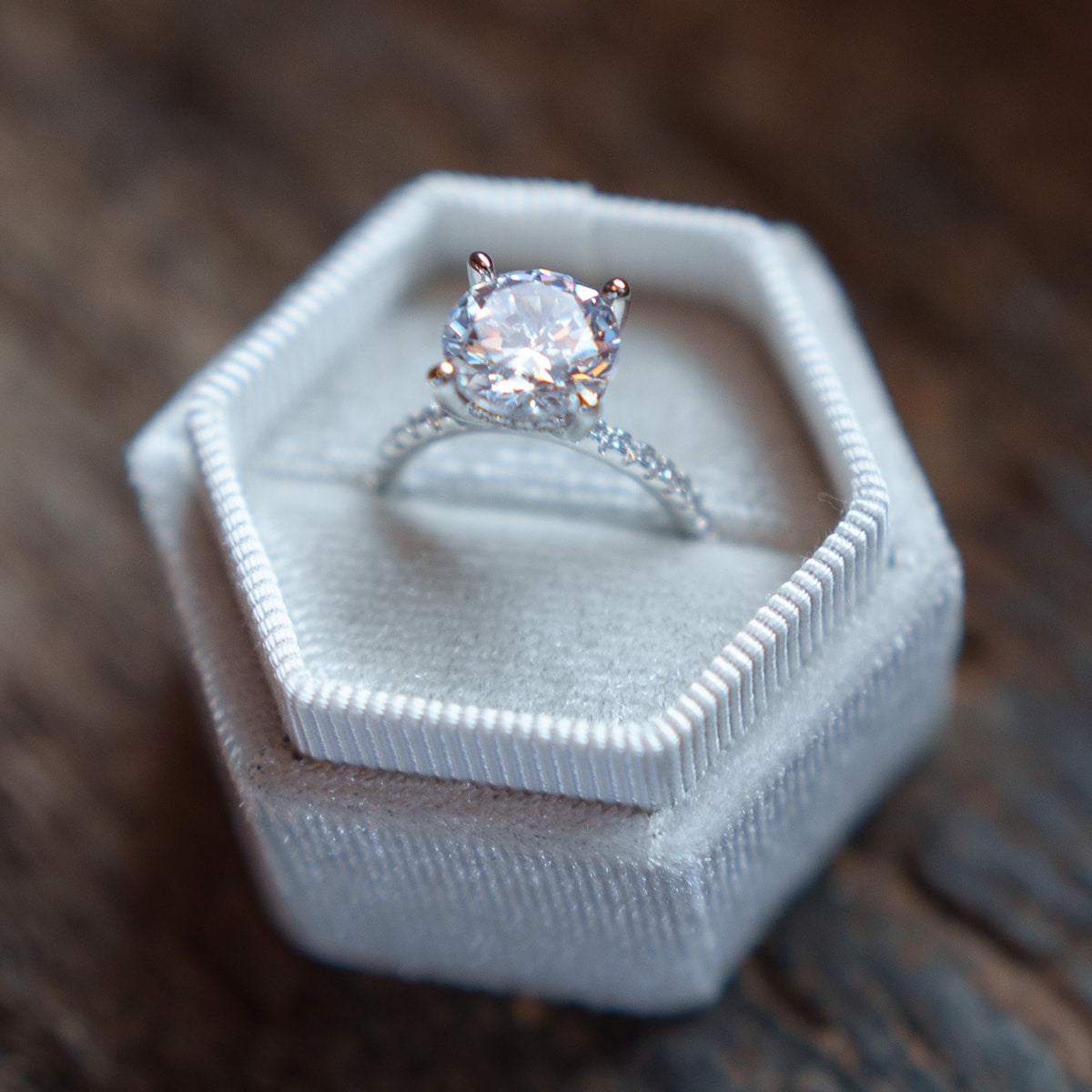 A silver solitaire engagement ring with a round clear gemstone, displayed in a white hexagonal velvet ring box on a rustic dark wood surface.