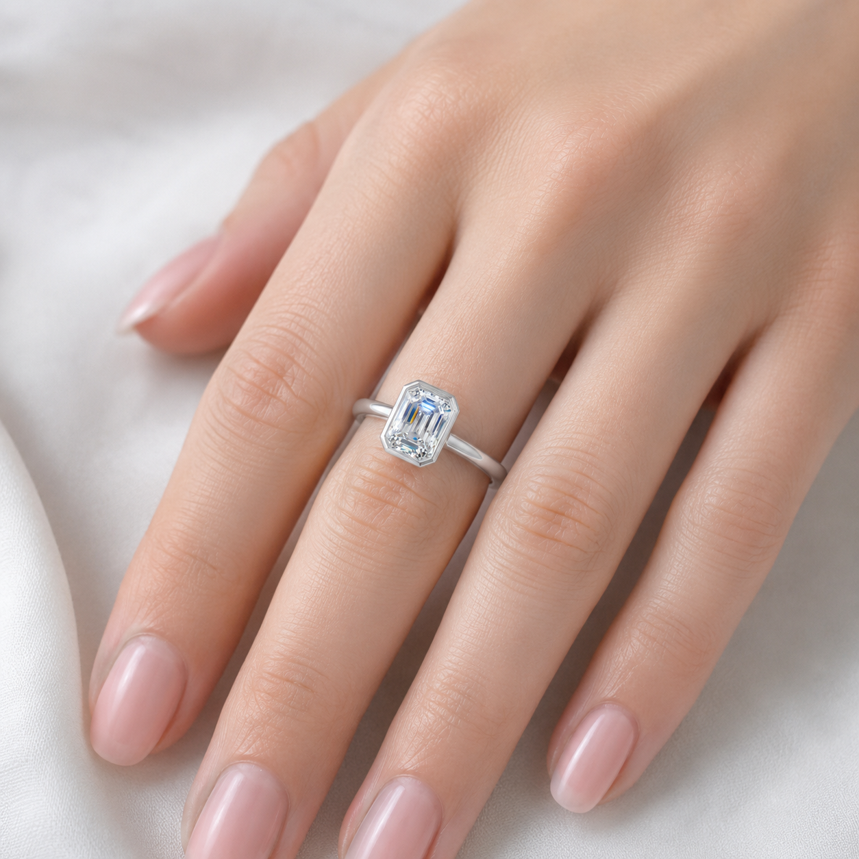 A silver ring with an emerald-cut clear gemstone worn on a woman's ring finger, shown against a soft white fabric background.