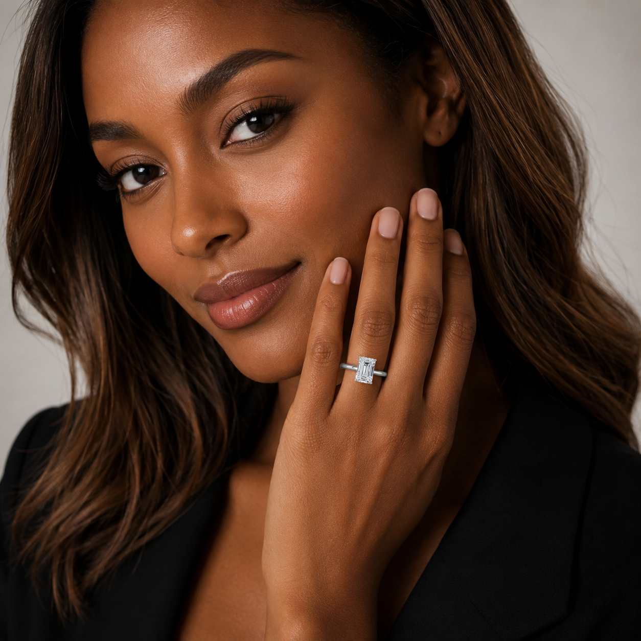 A close-up lifestyle shot of a woman wearing a silver engagement ring with a rectangular clear gemstone on her finger, posing with her hand near her face against a neutral light gray background.