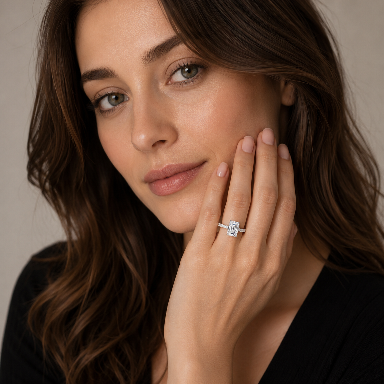 A close-up lifestyle shot of a woman with long brown hair wearing a silver ring with a rectangular clear gemstone on her finger, posing with her hand near her face against a soft beige background.