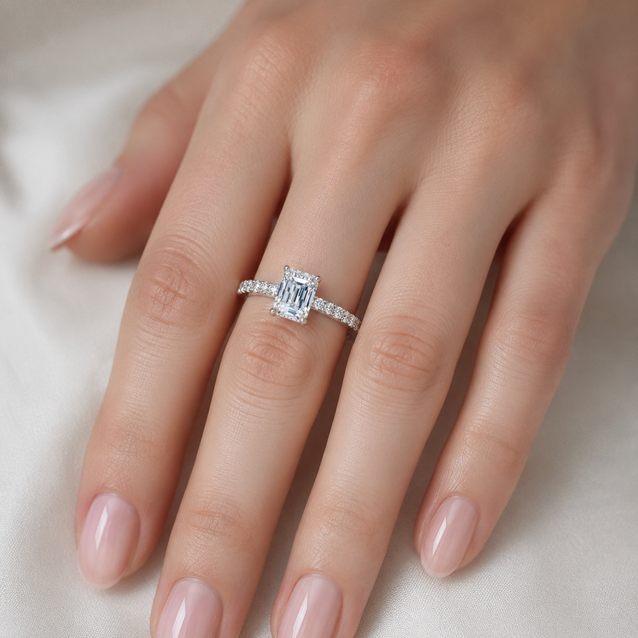 A close-up of a hand wearing a silver engagement ring with a rectangular emerald-cut diamond and small side stones, set against a soft white fabric background.