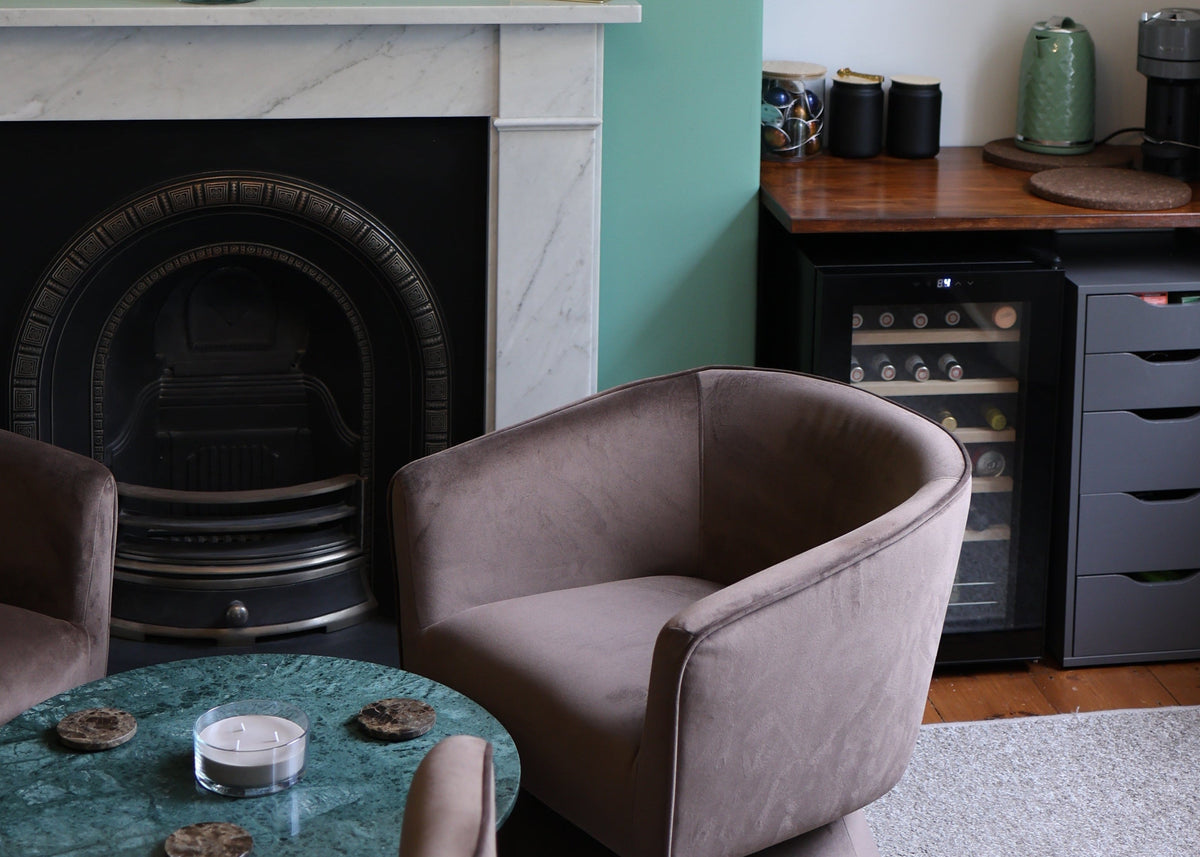 A modern living room corner featuring a teal-green wall, a white marble fireplace, two plush taupe armchairs, a round green marble coffee table, and a black wine refrigerator under a wood counter.