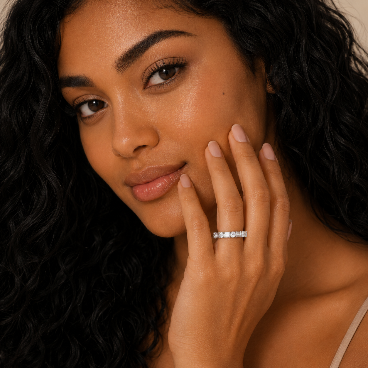 A close-up lifestyle shot of a woman with long, wavy black hair wearing a silver ring with small clear gemstones on her ring finger. She is touching her cheek with her hand, and the background is a soft, warm beige.