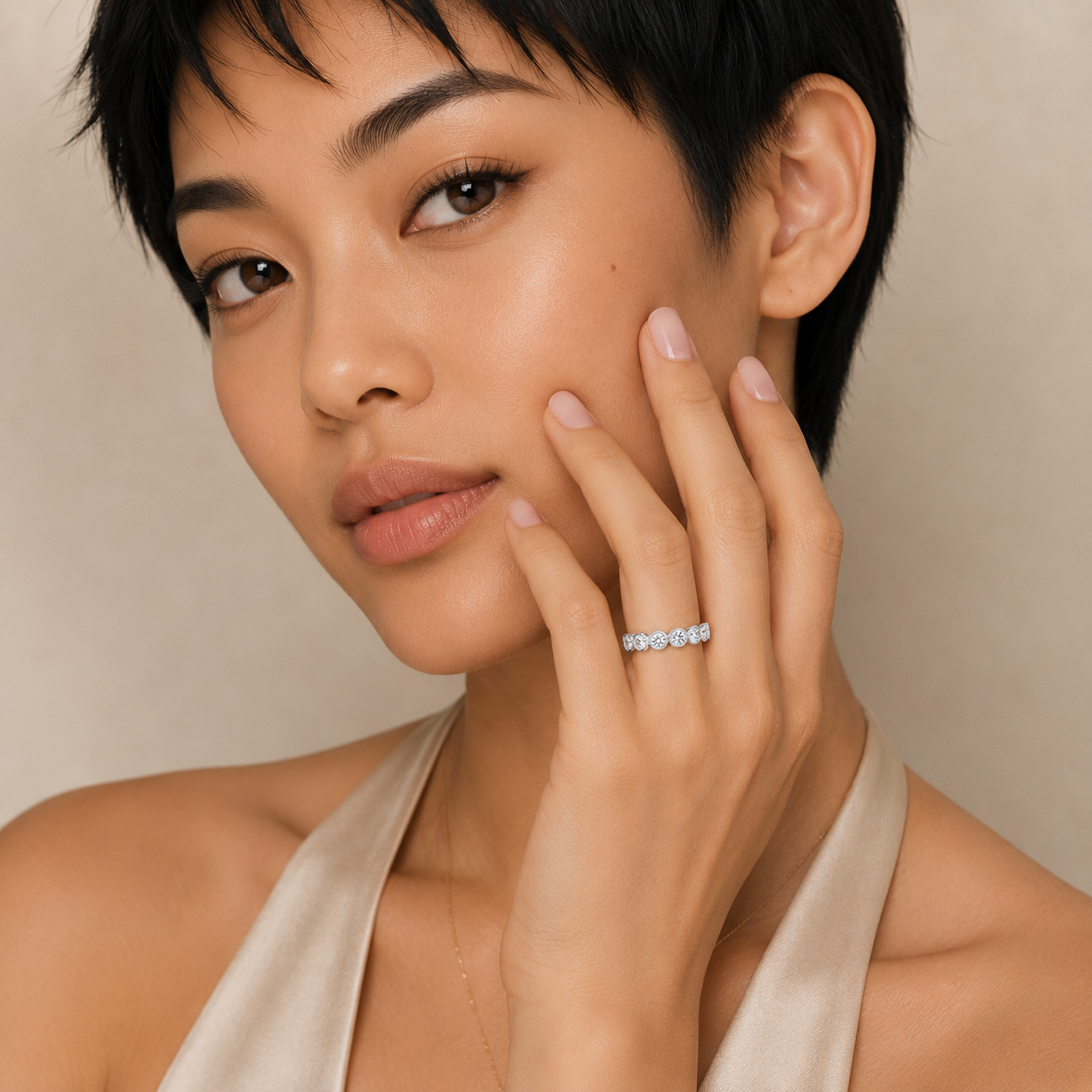 A close-up portrait of a woman with short dark hair wearing a delicate silver ring set with small clear stones on her ring finger, against a soft beige background.